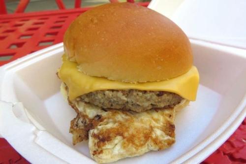 A sandwich with a golden bun, melted cheese, and a sausage patty on a bed of fried egg, served in a white container.