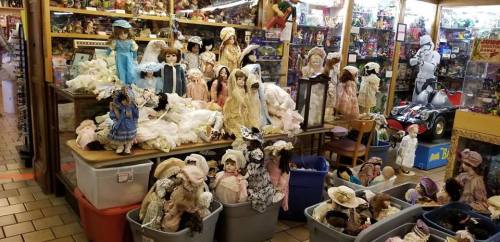 A collection of vintage dolls displayed on tables and in bins, surrounded by various toys and collectibles in a shop.