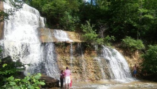 A scenic waterfall surrounded by lush greenery, with two people standing near the water's edge.