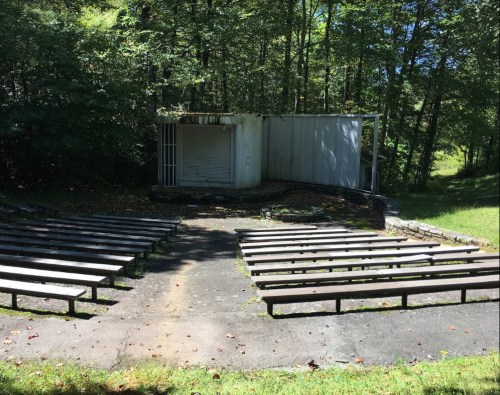 An outdoor amphitheater with wooden benches facing a small stage, surrounded by trees and greenery.