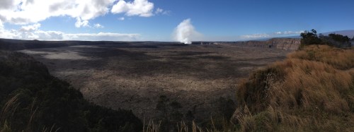A panoramic view of a volcanic crater with steam rising in the distance, surrounded by grass and blue sky.