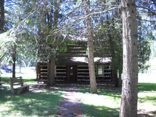 A rustic log cabin surrounded by trees, with a path leading to the entrance and a bench nearby.