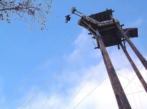 A person is zip-lining from a tall wooden tower against a blue sky with a few clouds.