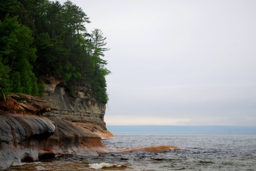 A rocky shoreline with lush green trees and a calm body of water under a cloudy sky.
