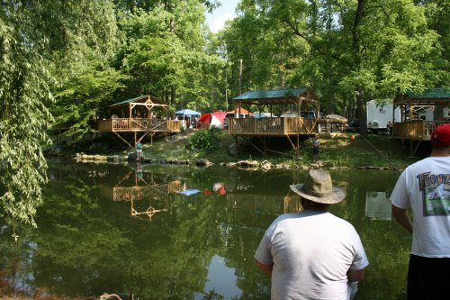 A serene pond surrounded by trees, with wooden gazebos and tents, and a person fishing in the foreground.