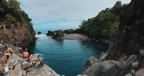 A scenic view of a rocky coastline with people relaxing by the water, surrounded by lush greenery and calm blue waters.