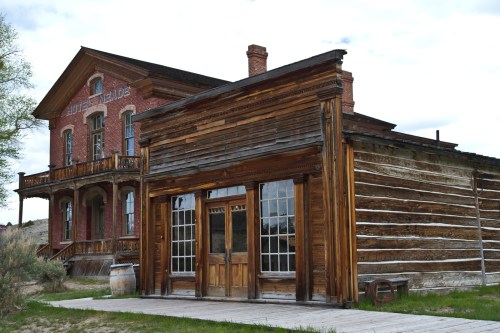 A rustic wooden building with large windows, set against a backdrop of a historic brick structure and cloudy sky.