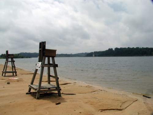 Lifeguard stands on a sandy beach by a calm lake under a cloudy sky. Trees line the opposite shore.