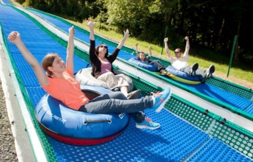 Four people on inflatable tubes, smiling and raising their arms, ready to slide down a blue, outdoor tubing slope.