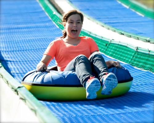 A girl joyfully sliding down a colorful water slide on a tube, wearing a bright orange shirt and jeans.