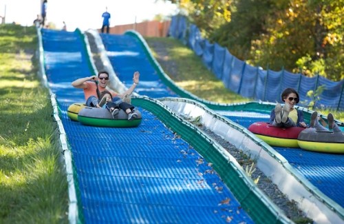 Two people joyfully sliding down a colorful tubing hill on a sunny day, surrounded by greenery and blue safety barriers.