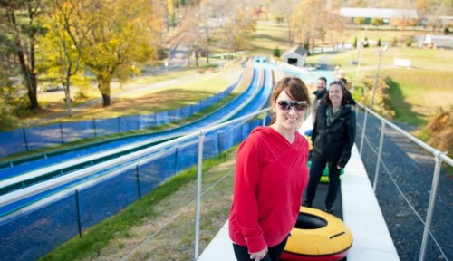 A group of people stands at the top of a colorful tubing slide, surrounded by trees and a sunny outdoor setting.