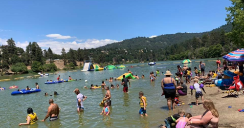 A busy lakeside scene with families enjoying water activities, inflatable structures, and a sunny mountain backdrop.