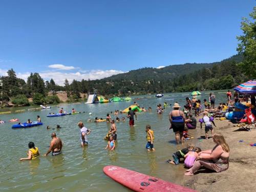 A busy lakeside scene with people swimming, floating on rafts, and relaxing on the shore under a clear blue sky.