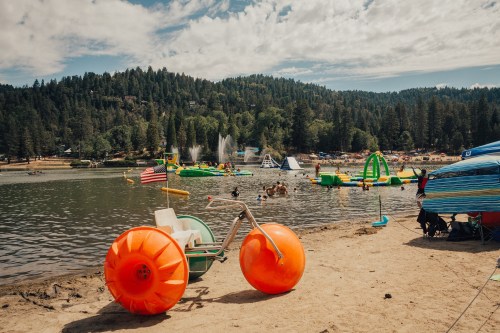 A sandy beach with colorful inflatable toys in the water, surrounded by trees and people enjoying a sunny day.