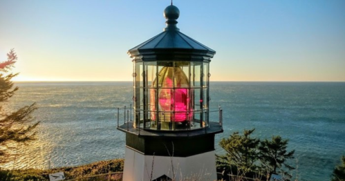 A lighthouse with a glass lantern sits by the ocean, illuminated by the sunset, surrounded by trees.