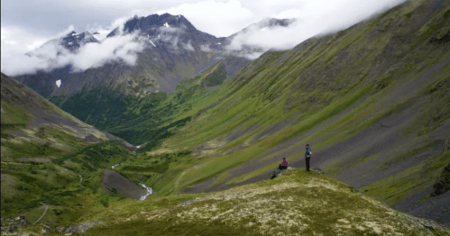 Two hikers stand on a grassy ridge overlooking a lush valley with mountains and clouds in the background.