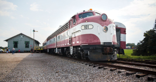 A vintage train in maroon and white parked beside a historic train station on a sunny day.