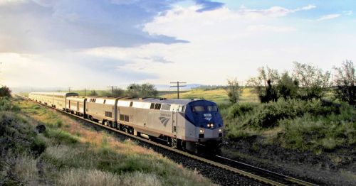 A train travels along a scenic route, surrounded by greenery and under a partly cloudy sky.