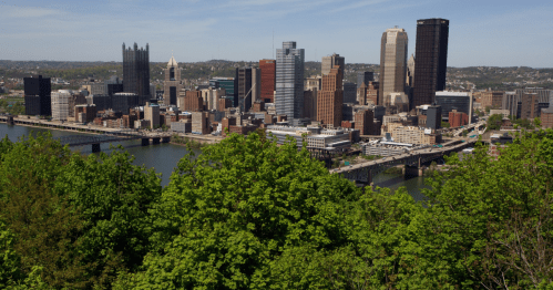 A panoramic view of Pittsburgh's skyline with green trees in the foreground and a river flowing through the city.
