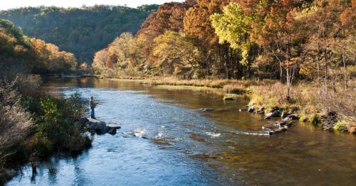 A serene river flows through a colorful autumn landscape, with a person fishing on a rocky bank.