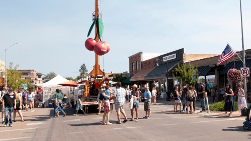 A lively street fair with people gathered, a cherry-themed decoration hanging from a crane, and vendor tents in the background.