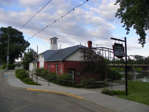 A red brick building with a gray roof, sign for the Chamber of Commerce, near a river and walking path.