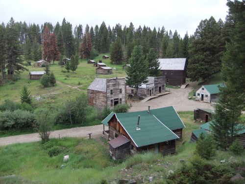 Aerial view of a rustic ghost town with wooden buildings surrounded by lush greenery and tall trees.
