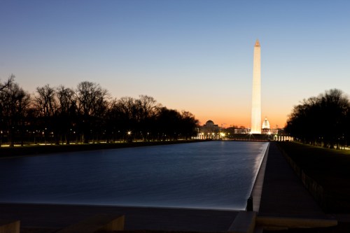 The Washington Monument illuminated at dusk, reflecting in the calm waters of the nearby pool.