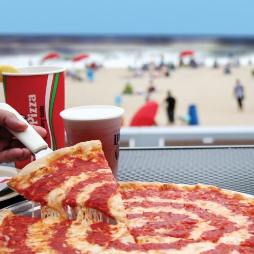 A hand lifts a slice of cheesy pizza from a table overlooking a beach with umbrellas and people in the background.