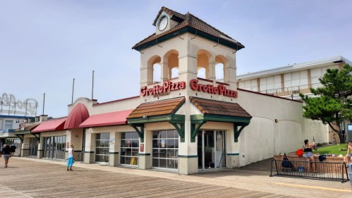 Grotto Pizza building on a boardwalk, featuring a bell tower and outdoor seating area. People walking nearby.