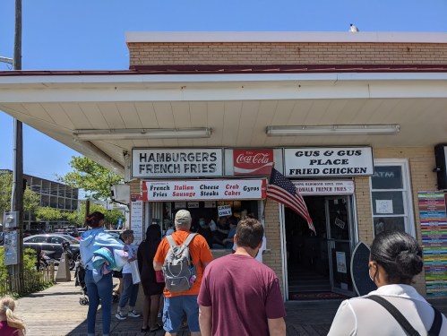 A busy food stand with signs for hamburgers, fries, and fried chicken, with a line of customers waiting outside.