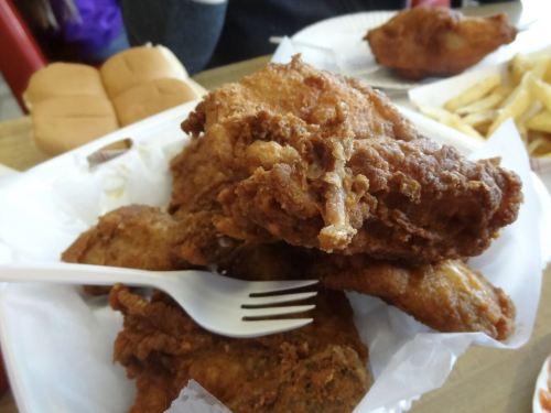 A plate of crispy fried chicken pieces with a plastic fork, accompanied by bread rolls and fries in the background.