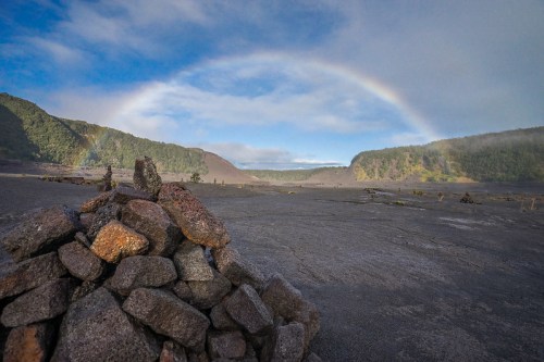 A rocky pile in the foreground with a rainbow arching over a volcanic landscape and lush green hills in the background.