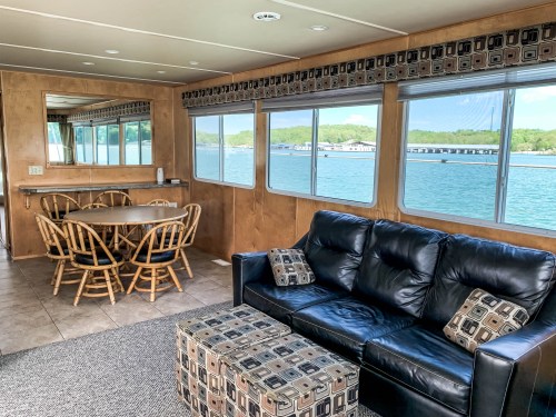 Interior of a houseboat featuring a dining area, a black sofa, and large windows overlooking the water.