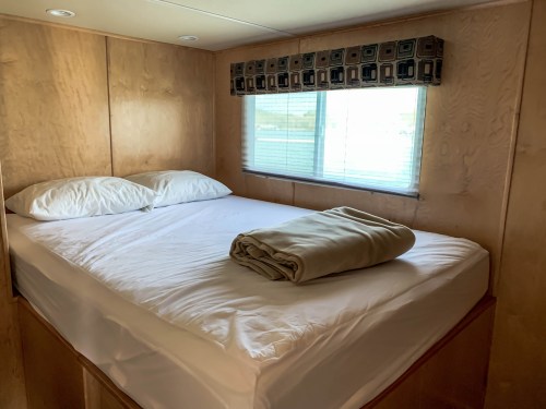 Cozy bedroom with a double bed, white linens, and a folded blanket, featuring wooden walls and a window with blinds.