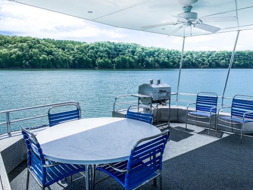 A spacious outdoor deck with a round table, blue chairs, and a grill, overlooking a serene lake and lush greenery.