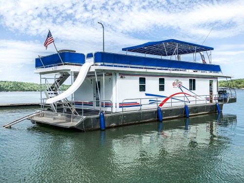 A colorful houseboat with a blue roof and slide, docked on calm water under a partly cloudy sky.