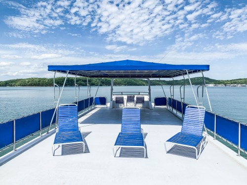 A spacious boat deck with blue lounge chairs under a canopy, overlooking calm water and a scenic landscape.