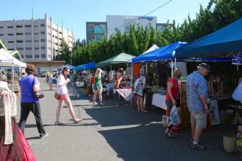 A bustling outdoor market with colorful tents, shoppers browsing various goods, and greenery in the background.