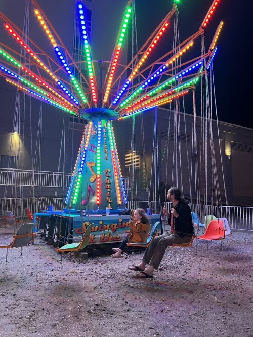A brightly lit swing ride at night, with colorful lights and two people sitting on swings, enjoying the atmosphere.