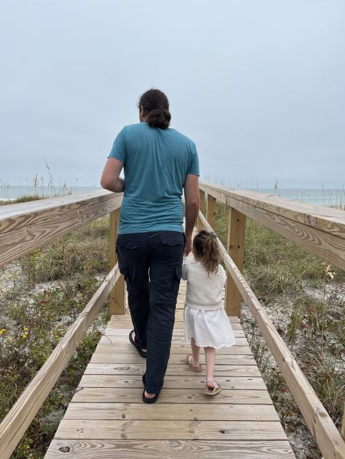 A man and a young girl walk hand in hand along a wooden path leading to the beach on a cloudy day.