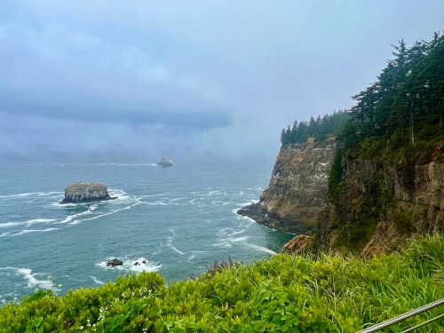 A misty coastal scene featuring cliffs, rocky outcrops, and waves crashing against the shore under a cloudy sky.