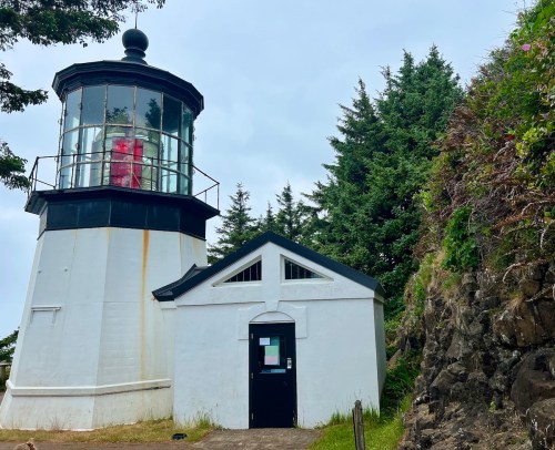 A white lighthouse with a black roof, surrounded by trees and rocky terrain, under a cloudy sky.