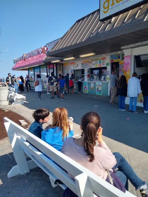 A busy ice cream shop with a long line, people enjoying ice cream, and a sunny day.