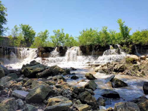 A serene waterfall cascades over rocks, surrounded by lush greenery under a clear blue sky.