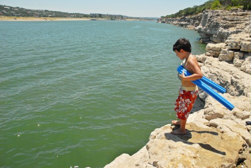 A boy in swim trunks stands on a rocky shore, holding pool noodles, looking at the water on a sunny day.