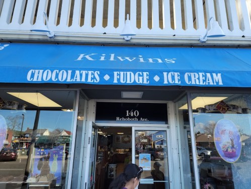 Storefront of Kilwins featuring a blue awning with the words "Chocolates, Fudge, Ice Cream" and the address 140 Rehoboth Ave.
