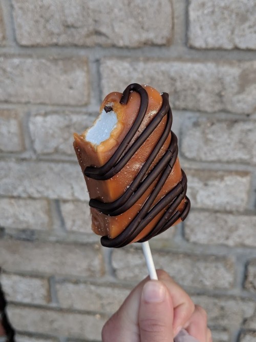 A partially eaten ice cream treat on a stick, coated in caramel and drizzled with chocolate, held against a brick wall.