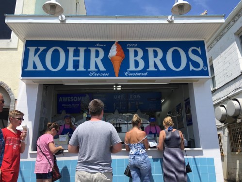 A busy ice cream stand with people ordering frozen custard on a sunny day. The sign reads "Kohr Bros Frozen Custard."